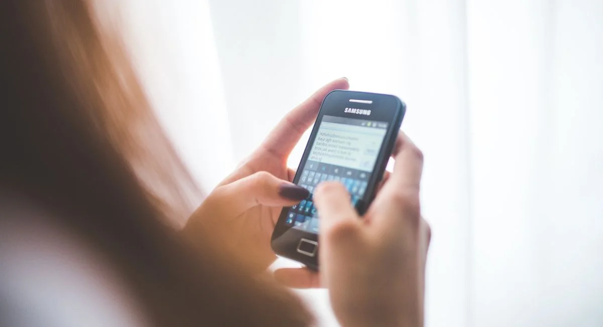 Close-up of a young woman typing on a Samsung smartphone at home. Bright indoor lighting.